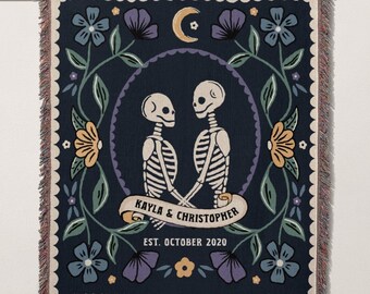 Puede incluir: Un tapiz tejido azul marino con un borde floral y dos esqueletos que se toman de la mano. El texto "KAYLA & CHRISTOPHER EST. OCTUBRE 2020" está impreso en el tapiz.