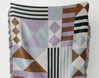 May include: A woven throw blanket with a geometric pattern in shades of brown, purple, blue, and white. The blanket has a fringe border.