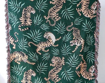 May include: A green woven throw blanket with a repeating pattern of tigers and palm leaves. The blanket has fringe on all sides.