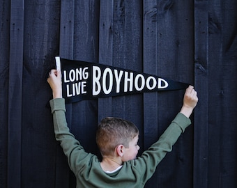 Puede incluir: Un niño sostiene una bandera negra con texto blanco que dice "Long Live Boyhood" sobre un fondo de madera oscura.