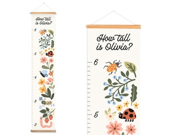 Puede incluir: Un medidor de altura crema con el texto "How tall is Olivia?" y un diseño floral e insectos. El medidor presenta una mariquita, una mariposa y flores. Las clavijas de madera en la parte superior e inferior miden hasta 183 cm.
