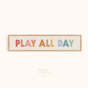 Puede incluir: Un letrero rectangular con marco de madera con las palabras "PLAY ALL DAY" en letras coloridas y redondeadas. Las letras son en tonos de rojo, rosa, naranja, amarillo, azul y verde. El fondo es de color beige claro.