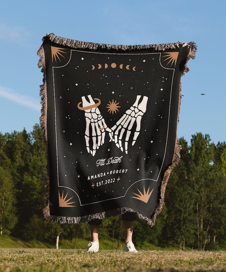 May include: Black woven throw blanket featuring a design of two skeleton hands holding each other, a sun, and a crescent moon. The text "Till Death" is written above the hands, and "Amanda + Robert Est. 2022" is written below the hands.