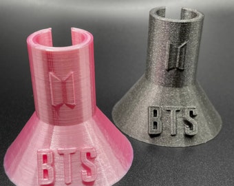 May include: Two 3D printed BTS phone stands, one pink and one black, with the BTS logo and the word "BTS" printed on the base.