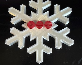 Può includere: Un ornamento a forma di fiocco di neve bianco a sei punte con una superficie strutturata e stratificata. Tre piccoli accenti circolari rossi sono posizionati al centro, ognuno con dettagli a linee orizzontali distinti.