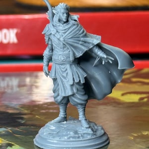 May include: A gray miniature figure of a fantasy character, possibly an elf, with a flowing cape and staff. The figure is standing on a circular base. The figure is wearing detailed armor and clothing.