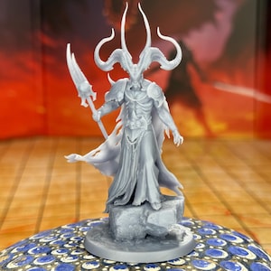 Può includere: Una statuetta grigia di un demone cornuto che tiene una lancia. La figura è su una base di pietra, con un mantello fluente e un'armatura. Lo sfondo è una scena rosso-arancio infuocata.