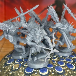 Puede incluir: Un grupo de figuras en miniatura grises, posiblemente para juegos de mesa, se disponen sobre una base decorativa. Las figuras, que parecen guerreros, sostienen una lanza o un arco. La base presenta un patrón azul y dorado, con un fondo degradado rojo y naranja.