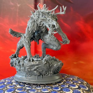 Puede incluir: Una figura gris detallada de una criatura con cuernos, pelaje y una cara esquelética, en una pose agachada. La figura está montada sobre una base texturizada. El fondo es un degradado de rojo y naranja.