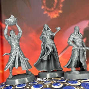 May include: Three gray miniature fantasy figures. One figure holds a bowl overhead, another holds a cup, and the third holds a curved blade. All are on round bases, with detailed clothing and facial features. The background is a blurred red and orange.