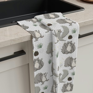 May include: A white kitchen towel with a gray squirrel pattern. The squirrels are depicted in a playful pose with their tails raised. The towel has a gray and white background with green pine needles and brown pine cones.