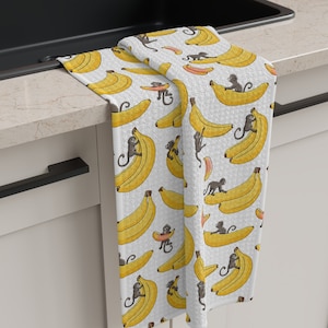 May include: A white kitchen towel with a pattern of yellow bananas and monkeys. The monkeys are holding bananas and riding on top of the bananas.