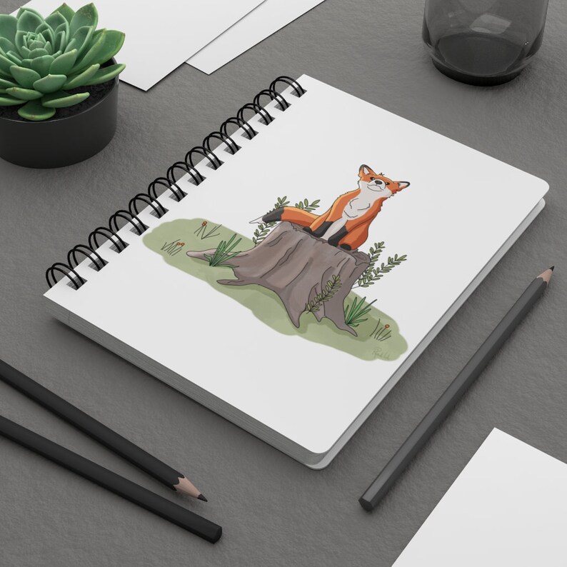 Op de afbeelding: Een wit spiraalnotitieblok met een cartoonillustratie van een oranje vos die op een boomstronk zit. De vos kijkt naar rechts en heeft een harige staart. De boomstronk is bruin en heeft groene bladeren eromheen.