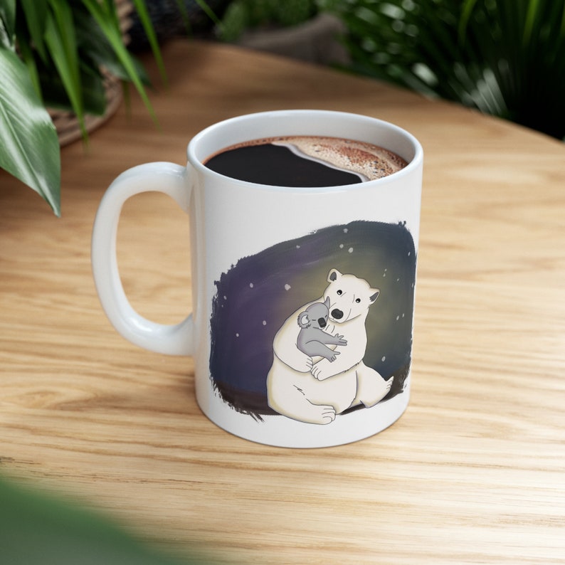 Pu&ograve; includere: Tazza in ceramica bianca con manico nero. La tazza presenta un orso polare bianco che abbraccia un koala grigio su uno sfondo blu scuro con stelle bianche.
