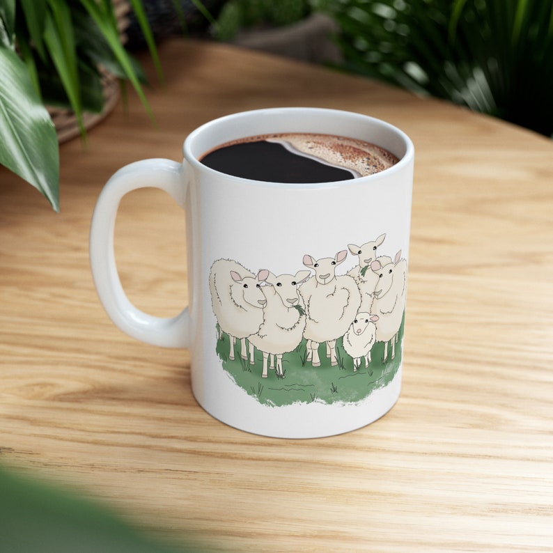 Op de afbeelding: Een witte keramische mok met een zwarte handgreep en een afbeelding van een groep schapen. De schapen zijn wit met zwarte ogen en neuzen en staan in een veld met groen gras.