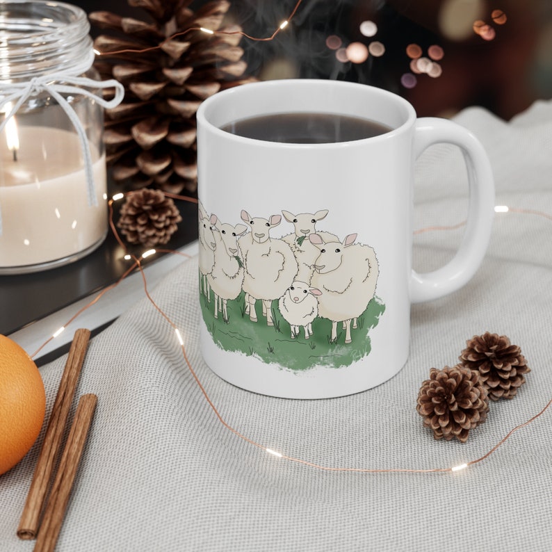 Op de afbeelding: Een witte keramische mok met een afbeelding van een groep schapen. De schapen zijn wit met zwarte ogen en neuzen. De mok heeft een zwart handvat en is gevuld met koffie.