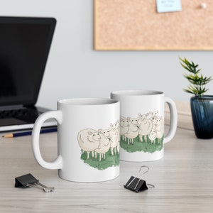 Op de afbeelding: Twee witte keramische mokken met een cartoonillustratie van een groep schapen. De schapen zijn wit met zwarte ogen en roze neuzen. De mokken staan op een licht houten oppervlak.