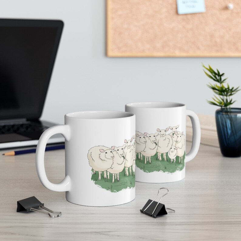Op de afbeelding: Twee witte keramische mokken met een cartoonillustratie van een groep schapen. De schapen zijn wit met zwarte ogen en roze neuzen. De mokken staan op een licht houten oppervlak.
