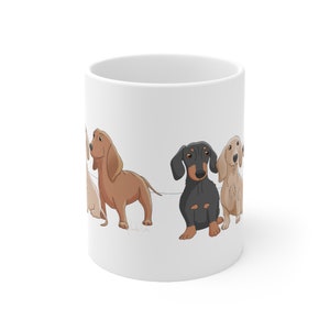May include: White ceramic mug with a cartoon illustration of four dachshund dogs. The dogs are brown, black, and cream colored. The dogs are all looking at the viewer.