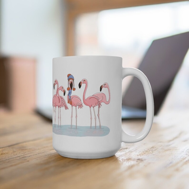 May include: White ceramic mug featuring a design of five pink flamingos, one sporting a blue scarf and a brown hat. The flamingos are standing in a line against a pale blue background.