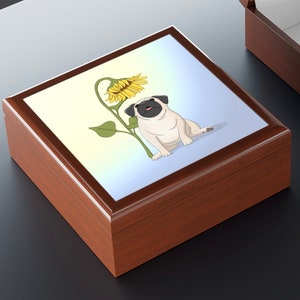 May include: A wooden jewelry box with a glossy finish. The lid features a cartoon illustration of a pug dog next to a sunflower. The background has a gradient of yellow and blue. The box is brown.
