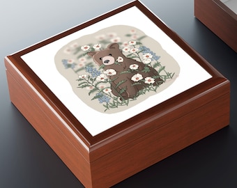 May include: A square wooden keepsake box with a dark brown finish and a hinged lid. The top of the lid features a square tile insert displaying a cartoon illustration of a brown bear sitting among white flowers and blue wildflowers on a beige background.