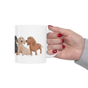 May include: White ceramic coffee mug with a cartoon illustration of four dachshund dogs in different colors. The dogs are lined up side by side, facing the viewer.