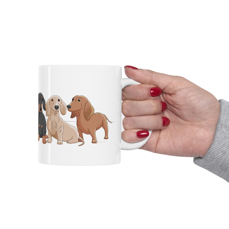 May include: White ceramic coffee mug with a cartoon illustration of four dachshund dogs in different colors. The dogs are lined up side by side, facing the viewer.