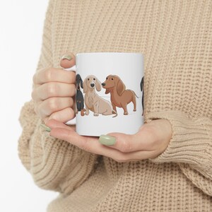May include: White ceramic mug with a colorful illustration of four dachshund dogs. The dogs are in a line, facing forward, and are all different colors. The mug is being held by a person's hands.