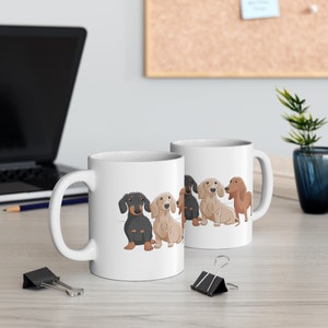 May include: White ceramic coffee mug with a design of four dachshund dogs in a row. The dogs are black, brown, and tan. The mug has a black handle.