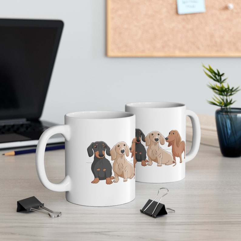 May include: White ceramic coffee mug with a design of four dachshund dogs in a row. The dogs are black, brown, and tan. The mug has a black handle.