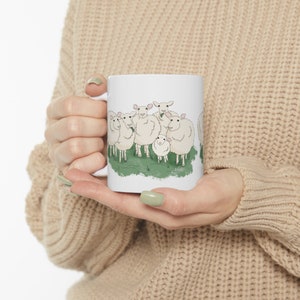 Op de afbeelding: Witte keramische mok met een cartoonillustratie van een groep schapen die in een veld met groen gras staan.