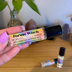Puede incluir: Un frasco de aceite esencial de Forest Witch con un rodillo y una etiqueta de colores arcoíris. La etiqueta dice "Forest Witch" en texto negro e incluye el texto "FOR GROUNDING ENERGIES & ENHANCING ONES CONNECTION TO MOTHER EARTH pine needles, crisp leaves, & dew drops HAND MADE WITH 100% NATURAL FRAGRANCE OILS".