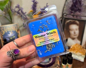 May include: A rectangular blue wax melt in a clear plastic clamshell container. The label reads Winter Witch, For Self Love & Emotional Stability, lavender and pine, Hand made in the USA with 100% natural fragrance oils. Dried lavender buds are embedded in the wax.