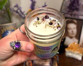 May include: A clear glass jar candle with white wax topped with small dark stones and dried herbs. The label reads FAMILIAR, CARAMELIZED BROWN SUGAR, RIPE FIG, AND RICH AMBER, THE WITCH'S CONCOCTION. The candle is held in a hand wearing a silver pentacle ring.