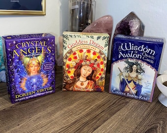 Könnte beinhalten: Drei Orakelkarten-Decks mit violetten, blauen und goldenen Akzenten. Die Decks sind betitelt mit "Crystal Angels Oracle Cards", "Goddess Dream Oracle" und "The Wisdom of Avalon Oracle Cards".