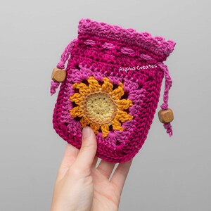 May include: A pink and yellow crocheted drawstring bag with a sunflower design. The bag has wooden beads on the drawstrings. The text "Alyssa Creates" is visible on the bag.