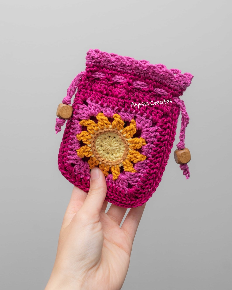 May include: A pink and yellow crocheted drawstring bag with a sunflower design. The bag has wooden beads on the drawstrings. The text "Alyssa Creates" is visible on the bag.