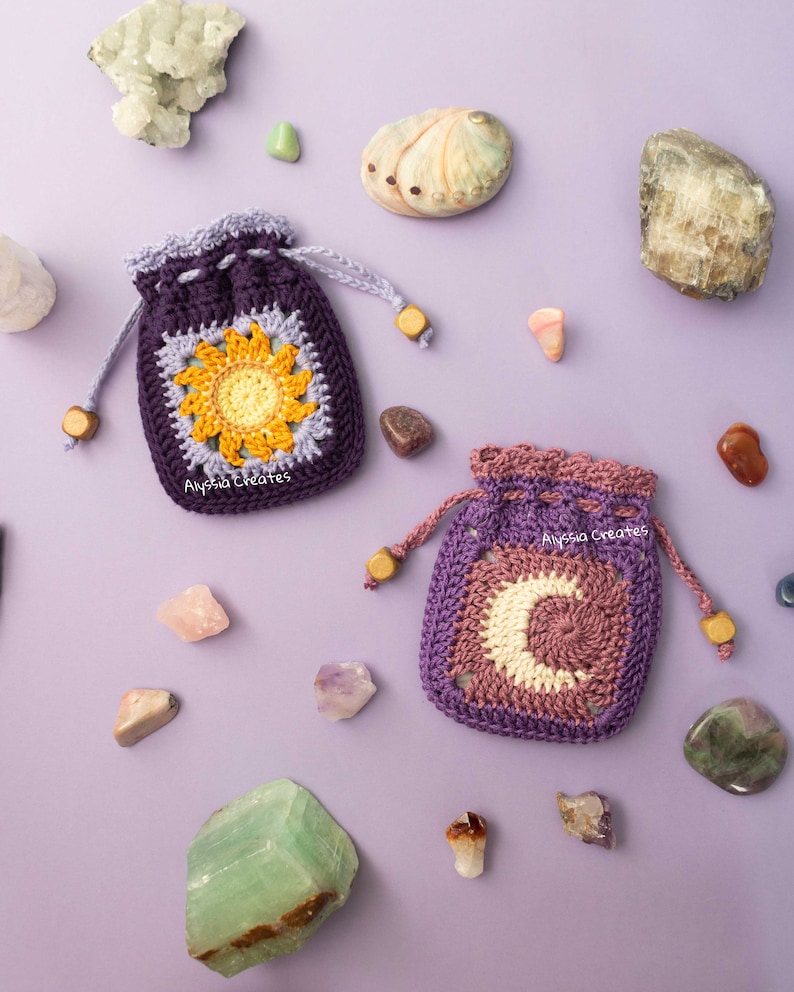 May include: Two crocheted pouches with a purple and lavender color scheme. One pouch features a yellow sunflower design and the other features a white crescent moon design. Both pouches are surrounded by various crystals and stones on a purple background. The text "Alyssa Creates" is visible on both pouches.