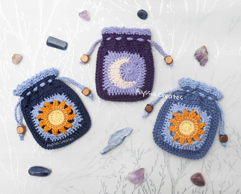 May include: Three crocheted drawstring pouches with different designs. The pouches are blue, purple, and light blue. The designs are a sun, a moon, and a flower. The pouches are on a white background with small stones.