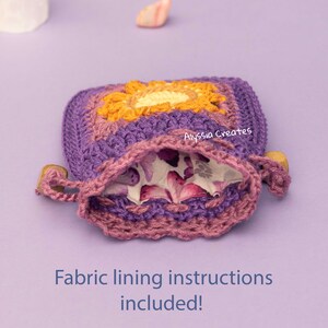 May include: Purple crochet pouch with a floral fabric lining and a wooden toggle closure. The pouch has a yellow and orange granny square design. "Alyssa Creates" is written on the pouch. "Fabric lining instructions included!" is written below the pouch.