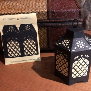 May include: A black paper lantern with intricate cut-out designs and a white interior. The lantern is on a wooden surface with a piece of paper that says "www.witchnextdoorstore.com".