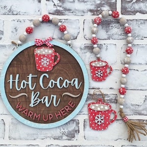 May include: A wooden sign with a blue border and the text "Hot Cocoa Bar Warm Up Here" in white. The sign is decorated with a red and white polka dot mug with marshmallows and a snowflake. The sign is hanging from a white and red beaded garland.