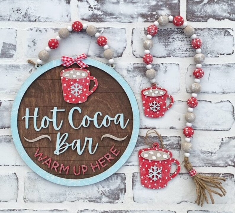 May include: A wooden sign with a blue border and the text "Hot Cocoa Bar Warm Up Here" in white. The sign is decorated with a red and white polka dot mug with marshmallows and a snowflake. The sign is hanging from a white and red beaded garland.