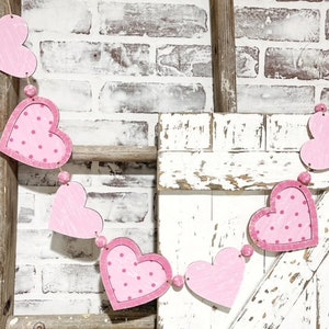K&ouml;nnte beinhalten: Eine Girlande aus rosa und wei&szlig;en Herzen mit Punkten h&auml;ngt an einer wei&szlig; gestrichenen Holzleiter. Die Girlande ist aus Filz oder Stoff und perfekt f&uuml;r die Valentinstagsdekoration.