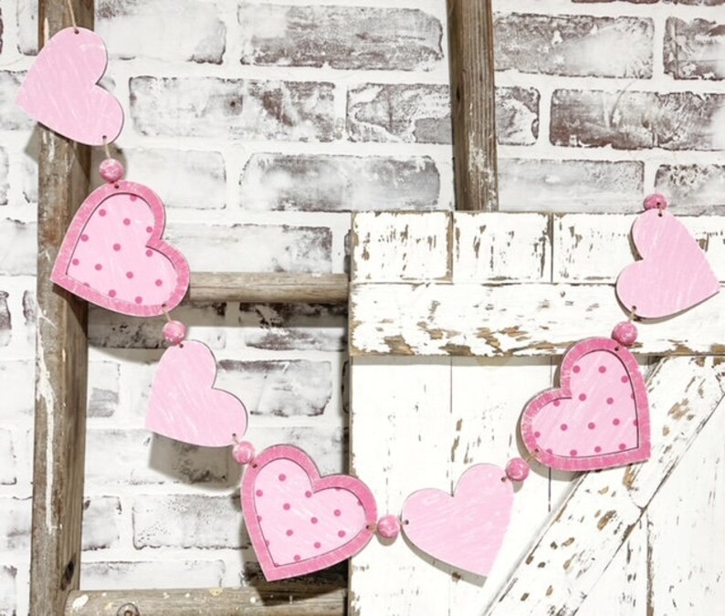 K&ouml;nnte beinhalten: Eine Girlande aus rosa und wei&szlig;en Herzen mit Punkten h&auml;ngt an einer wei&szlig; gestrichenen Holzleiter. Die Girlande ist aus Filz oder Stoff und perfekt f&uuml;r die Valentinstagsdekoration.