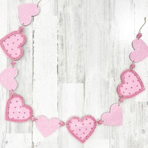 K&ouml;nnte beinhalten: Eine rosa-wei&szlig;e Herzgirlande mit rosa Punkten. Die Girlande ist aus Filz und mit einer Schnur zusammengef&uuml;gt. Die Girlande h&auml;ngt an einem wei&szlig;en Holzhintergrund.