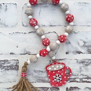 May include: A red and white wooden bead garland with a red and white mug charm. The mug has a snowflake and white dots. The garland has a brown and white tassel.