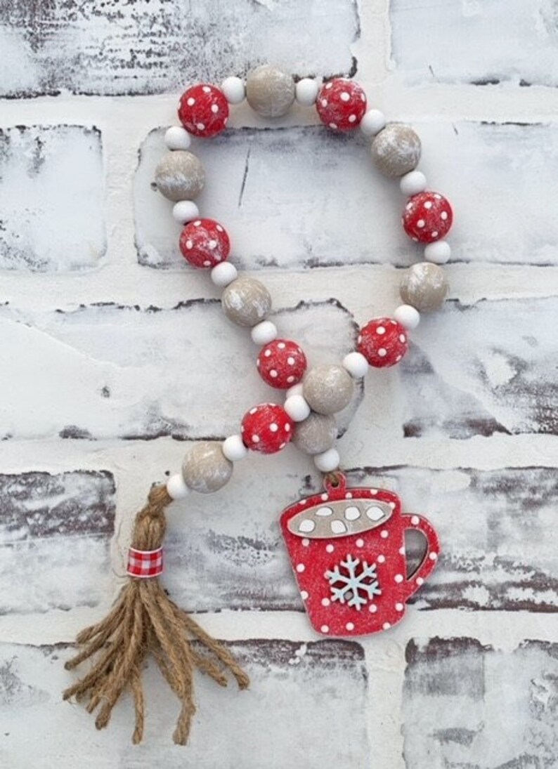 May include: A red and white wooden bead garland with a red and white mug charm. The mug has a snowflake and white dots. The garland has a brown and white tassel.