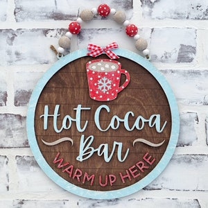 May include: A round wooden sign with a blue border and the text "Hot Cocoa Bar Warm Up Here". The sign has a red and white mug with marshmallows and a snowflake on it. The sign is decorated with a red and white gingham bow and a string of white and red beads.
